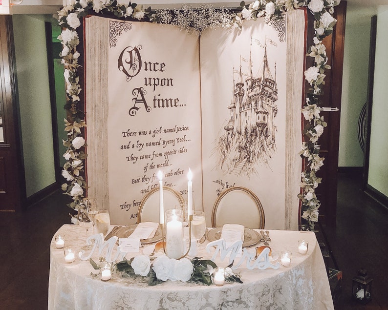 Puede incluir: Una mesa de boda con un mantel blanco, flores blancas y velas. El tel&oacute;n de fondo es un gran libro abierto con el texto "&Eacute;rase una vez..." y un dibujo de un castillo.