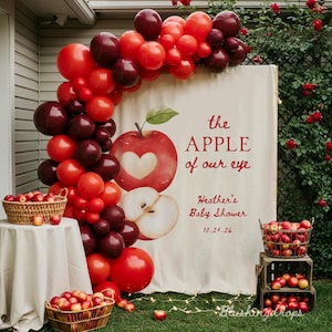 May include: A baby shower display featuring a red and burgundy balloon arch. A banner with an apple design and the text "the APPLE of our eye" is visible. Baskets of apples and a table with a white cloth are also present.