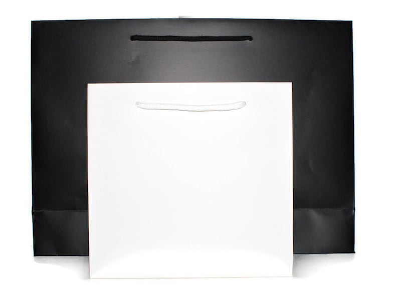 May include: Two paper gift bags, one black and one white, with handles. The bags are sitting on a white surface.