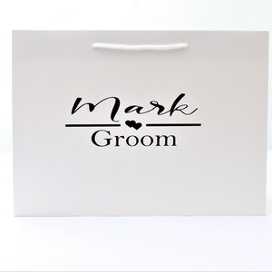 May include: White gift bag with black text that reads "Mark Groom" with two small hearts above the word "Groom".