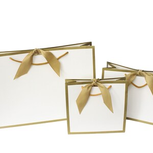 Puede incluir: Tres bolsas de regalo blancas de diferentes tama&ntilde;os, cada una con un borde dorado met&aacute;lico en la parte superior y los lados. Cada bolsa est&aacute; adornada con una cinta de sat&eacute;n dorada atada en un lazo en el centro, complementada con asas de cord&oacute;n dorado fino.