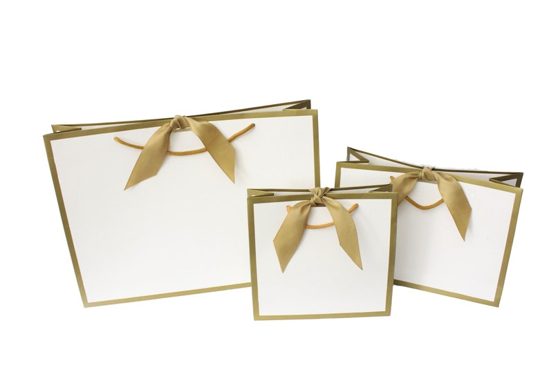 Puede incluir: Tres bolsas de regalo blancas de diferentes tama&ntilde;os, cada una con un borde dorado met&aacute;lico en la parte superior y los lados. Cada bolsa est&aacute; adornada con una cinta de sat&eacute;n dorada atada en un lazo en el centro, complementada con asas de cord&oacute;n dorado fino.