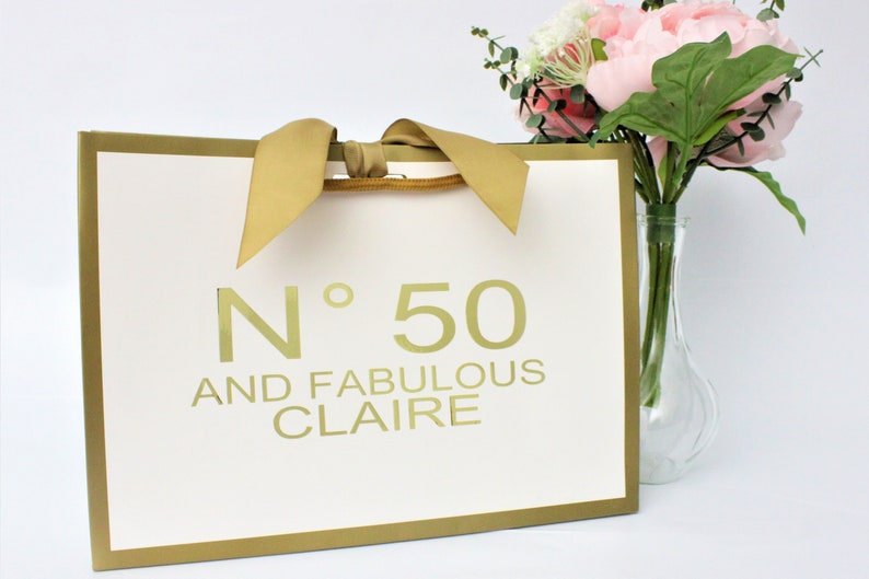 Puede incluir: Una bolsa de regalo rectangular blanca con borde dorado y asas de cinta dorada. El frente presenta el texto dorado N&deg; 50 AND FABULOUS CLAIRE. Un jarr&oacute;n de vidrio transparente con peon&iacute;as rosas y hojas de eucalipto se encuentra junto a la bolsa.