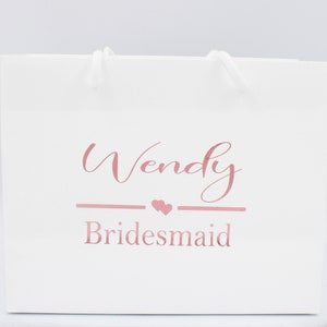 May include: White gift bag with a pink inscription that reads "Wendy Bridesmaid" with two small pink hearts.