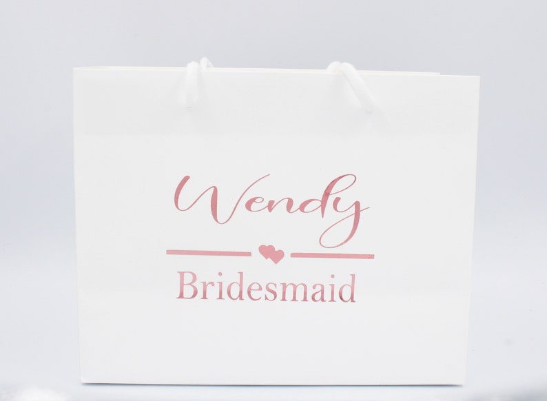 May include: White gift bag with a pink inscription that reads "Wendy Bridesmaid" with two small pink hearts.