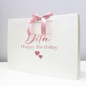 May include: White gift bag with a pink ribbon and the text "Dita Happy Birthday" in pink lettering with two pink hearts.