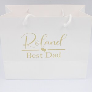 May include: White gift bag with gold lettering that says "Roland Best Dad" with two small hearts above the word "Best".
