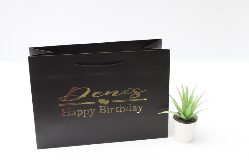 May include: Black gift bag with a gold inscription that reads "Denis Happy Birthday".