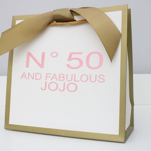Puede incluir: Una bolsa de regalo blanca con bordes met&aacute;licos dorados y un lazo de cinta dorada en la parte superior. El frente presenta el texto rosa N&deg; 50 AND FABULOUS JOJO impreso en una fuente sans-serif. La bolsa tiene una forma cuadrada con base plana.