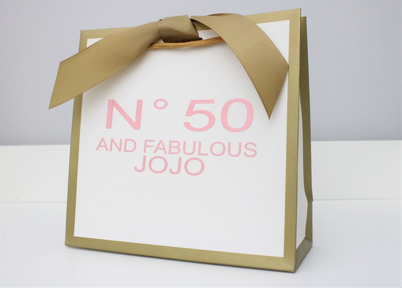 Puede incluir: Una bolsa de regalo blanca con bordes met&aacute;licos dorados y un lazo de cinta dorada en la parte superior. El frente presenta el texto rosa N&deg; 50 AND FABULOUS JOJO impreso en una fuente sans-serif. La bolsa tiene una forma cuadrada con base plana.