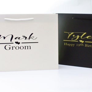 May include: Two white and black gift bags with personalized text. The white bag has black text that says "Mark Groom" with two small hearts. The black bag has gold text that says "Tyler Happy 18th Birthday" with two small hearts.
