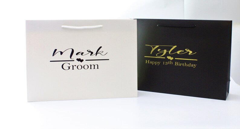 May include: Two white and black gift bags with personalized text. The white bag has black text that says "Mark Groom" with two small hearts. The black bag has gold text that says "Tyler Happy 18th Birthday" with two small hearts.