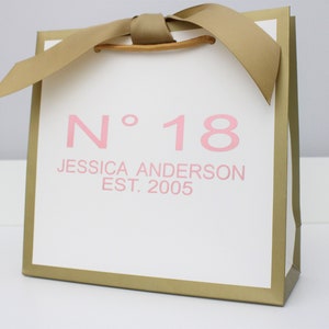 Puede incluir: Una bolsa de regalo blanca con bordes met&aacute;licos dorados y un asa de cinta dorada. El frente presenta texto rosa que dice N&deg; 18, JESSICA ANDERSON y EST. 2005. La bolsa tiene una forma cuadrada y un dise&ntilde;o estructurado con un acabado liso.