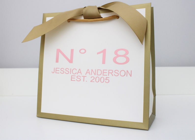 Puede incluir: Una bolsa de regalo blanca con bordes met&aacute;licos dorados y un asa de cinta dorada. El frente presenta texto rosa que dice N&deg; 18, JESSICA ANDERSON y EST. 2005. La bolsa tiene una forma cuadrada y un dise&ntilde;o estructurado con un acabado liso.