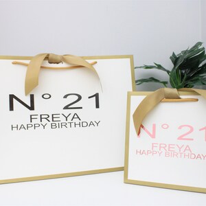 Puede incluir: Dos bolsas de regalo blancas con borde dorado y asas de cinta dorada. La bolsa m&aacute;s grande tiene texto negro que dice N&deg; 21 FREYA HAPPY BIRTHDAY. La bolsa m&aacute;s peque&ntilde;a tiene texto rosa que dice N&deg; 21 FREYA HAPPY BIRTHDAY. Ambas son rectangulares.