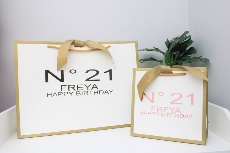 Puede incluir: Dos bolsas de regalo blancas con borde dorado y asas de cinta dorada. La bolsa m&aacute;s grande tiene texto negro que dice N&deg; 21 FREYA HAPPY BIRTHDAY. La bolsa m&aacute;s peque&ntilde;a tiene texto rosa que dice N&deg; 21 FREYA HAPPY BIRTHDAY. Ambas son rectangulares.