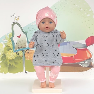 Puede incluir: Un conjunto de muñeca gris y rosa con estampado de panda y un gorro rosa con estampado de conejo. La muñeca lleva un jersey gris con capucha con estampado de panda y un pantalón rosa con estampado de conejo. La muñeca lleva un gorro rosa con estampado de conejo.