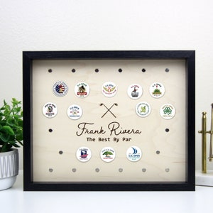 May include: A black framed shadow box with a wood background. The shadow box has a collection of golf ball markers with the US Open logo. The shadow box also has a personalized inscription that reads "Frank Rivera The Best By Par".