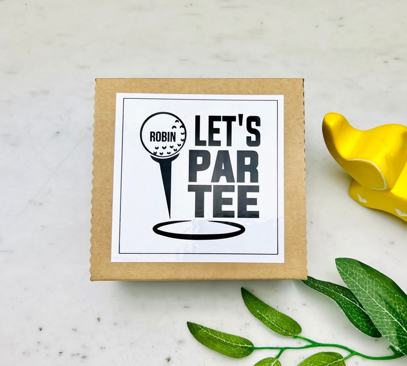 May include: A square, corrugated cardboard box with a white label featuring a golf ball on a tee and the words "LET'S PAR TEE" in bold, black letters. The name "ROBIN" is above the golf ball. The box is on a white surface.