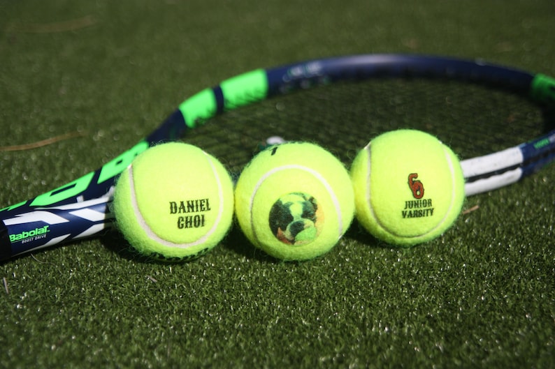 Pu&ograve; includere: Tre palline da tennis gialle con testo diverso stampato su ciascuna. La pallina a sinistra dice "Daniel Choi", la pallina al centro ha un'immagine di un cane e la pallina a destra dice "6 Junior Varsity".