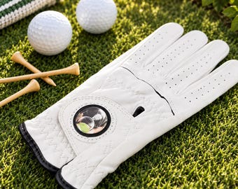 May include: A white golf glove with perforated fingers and a circular black-rimmed magnetic ball marker on the wrist strap. The glove features a textured palm area and black trim along the edges, positioned alongside two white golf balls and tees.
