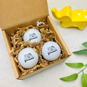 May include: A set of three white golf balls in a cardboard box, nestled in brown paper filler. Each ball has the text "IT'S TIME TO SUIT UP, WILL YOU BE MY Groomsman?" printed in black. A yellow elephant figurine is in the background.