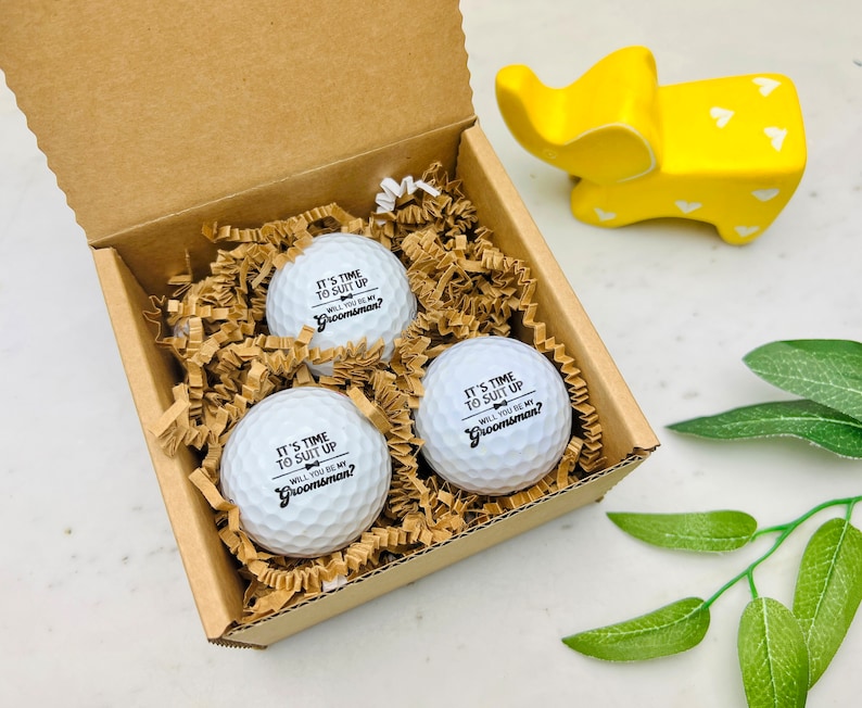 May include: A set of three white golf balls in a cardboard box, nestled in brown paper filler. Each ball has the text "IT'S TIME TO SUIT UP, WILL YOU BE MY Groomsman?" printed in black. A yellow elephant figurine is in the background.