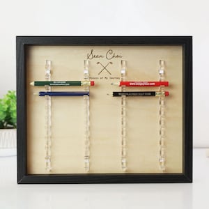 May include: A framed display with pencils, featuring the name "Sean Choi" and the text "Places of My Journey." The frame holds pencils in green, blue, red, and black, each with printed text. Clear acrylic holders suspend the pencils.