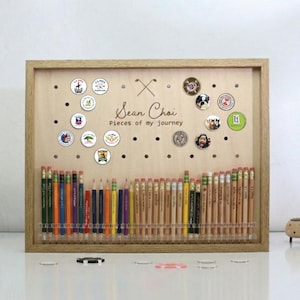 May include: A wooden framed display board with pencils and golf ball markers. The board has the text "Sean Choi Pieces of my journey". The pencils are arranged in a row below the markers.