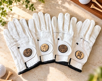 May include: Four white leather golf gloves with black trim and perforated fingers. Each glove features a circular wooden patch on the wrist strap, engraved with unique monograms including E, SH, MJR, and TH, alongside golf-themed icons.