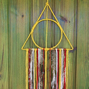 May include: A handmade wall hanging featuring a yellow triangle and circle design. Below, strands of yellow, red, white, and brown yarn hang down. Small metal charms are attached to some of the strands. The background is a green wooden surface.