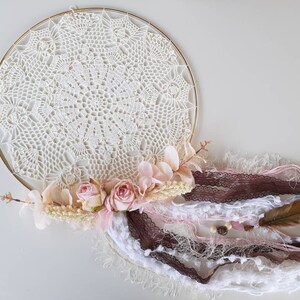 May include: A white crocheted dream catcher with a gold hoop, pink and white flowers, brown and white feathers, and white and pink ribbons.