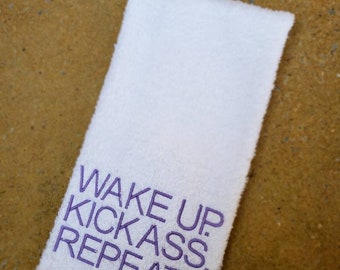 以下が含まれることがあります： 紫色の刺繍で「Wake Up. Kickass. Repeat.」と書かれた白いハンドタオルです。