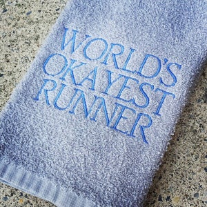 Puede incluir: Toalla gris con texto bordado en azul que dice "World's Okayest Runner".