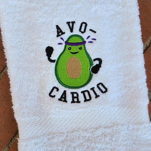 Pode incluir: Toalha branca com um abacate bordado usando uma faixa roxa e levantando pesos. O texto "AVO-CARDIO" está bordado abaixo do abacate.