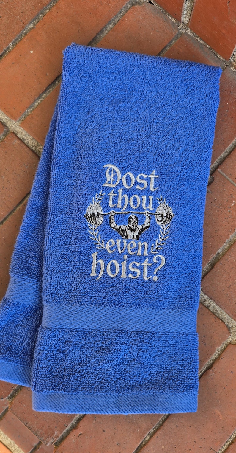 Puede incluir: Toalla de gimnasio azul con la frase bordada "Dost thou even hoist?" y un dise&ntilde;o de halterofilia. La toalla doblada tiene una apariencia texturizada, ideal para el gimnasio.