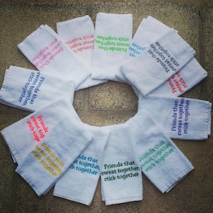 May include: Ten white towels with the phrase "Friends that sweat together, stick together" embroidered in different colors. The towels are arranged in a circle.