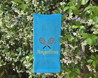Könnte beinhalten: Ein helles türkisfarbenes Handtuch mit einer Stickerei aus zwei gekreuzten schwarzen Tennisschlägern. Unter den Schlägern ist der Name Angelina in einer goldenen Schreibschrift eingestickt. Das Handtuch hat eine weiche Frotteeoberfläche.