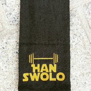 Może przedstawiać: Czarny ręcznik do ćwiczeń z haftem w kolorze złotym przedstawiającym sztangę i napis "HAN SWOLO".