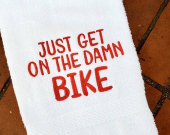 May include: White towel with red embroidered text that reads "Just get on the damn bike".