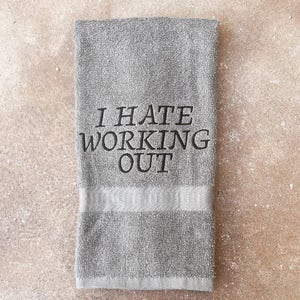 May include: A gray, rectangular towel with the embroidered text "I HATE WORKING OUT" in black. The towel has a lighter gray band near the bottom. The towel is made of a soft, absorbent material.