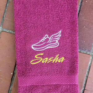 Puede incluir: Una toalla de mano color magenta con un bordado blanco que muestra el contorno de una zapatilla alada sobre el nombre Sasha, cosido en letra cursiva amarilla. La toalla tiene una superficie de rizo texturizada y un borde tejido decorativo.