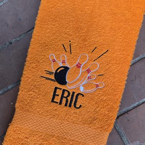 以下が含まれることがあります： オレンジ色のタオルに、ボウリングピンの刺繍と黒い文字で「ERIC」と刺繍されています。