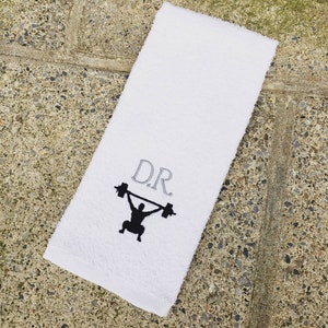 Puede incluir: Toalla de algodón blanca con las iniciales "D.R." bordadas en gris y una silueta negra de una persona levantando una barra.