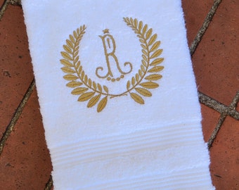 May include: White hand towel with a gold embroidered laurel wreath and the letter "R". The towel has a textured surface and horizontal stripes near the bottom. A decorative and personalized item.