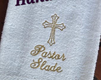 May include: White hand towel with gold embroidery of a cross and the text "Pastor Slade". The towel is 16x26 inches.