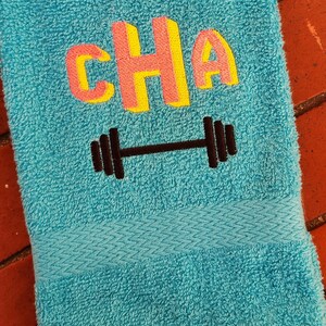 Puede incluir: Una toalla de color azul turquesa con la palabra "CHA" bordada en letras amarillas y rosas con un diseño de mancuerna negra debajo.