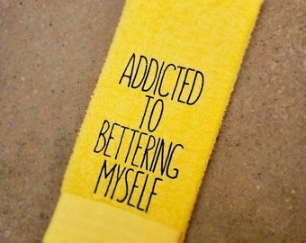 May include: A bright yellow towel with the text "ADDICTED TO BETTERING MYSELF" in black. The towel is rectangular with a folded bottom edge. The background is a neutral, textured surface.