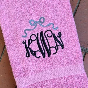 May include: A pink towel with a black embroidered monogram "K.L.N." with a silver bow above it.