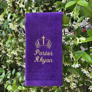 May include: A deep purple towel with gold embroidery. The design includes an open book, a cross, and the name "Pastor Rhyan" in a cursive font. The towel is hanging in front of a background of green leaves and white flowers.